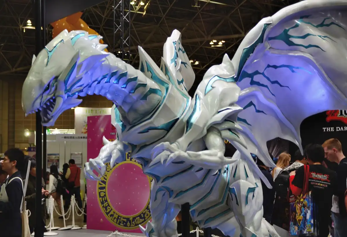 A large, white and blue dragon statue with glowing accents is displayed at a convention, surrounded by attendees. The text…