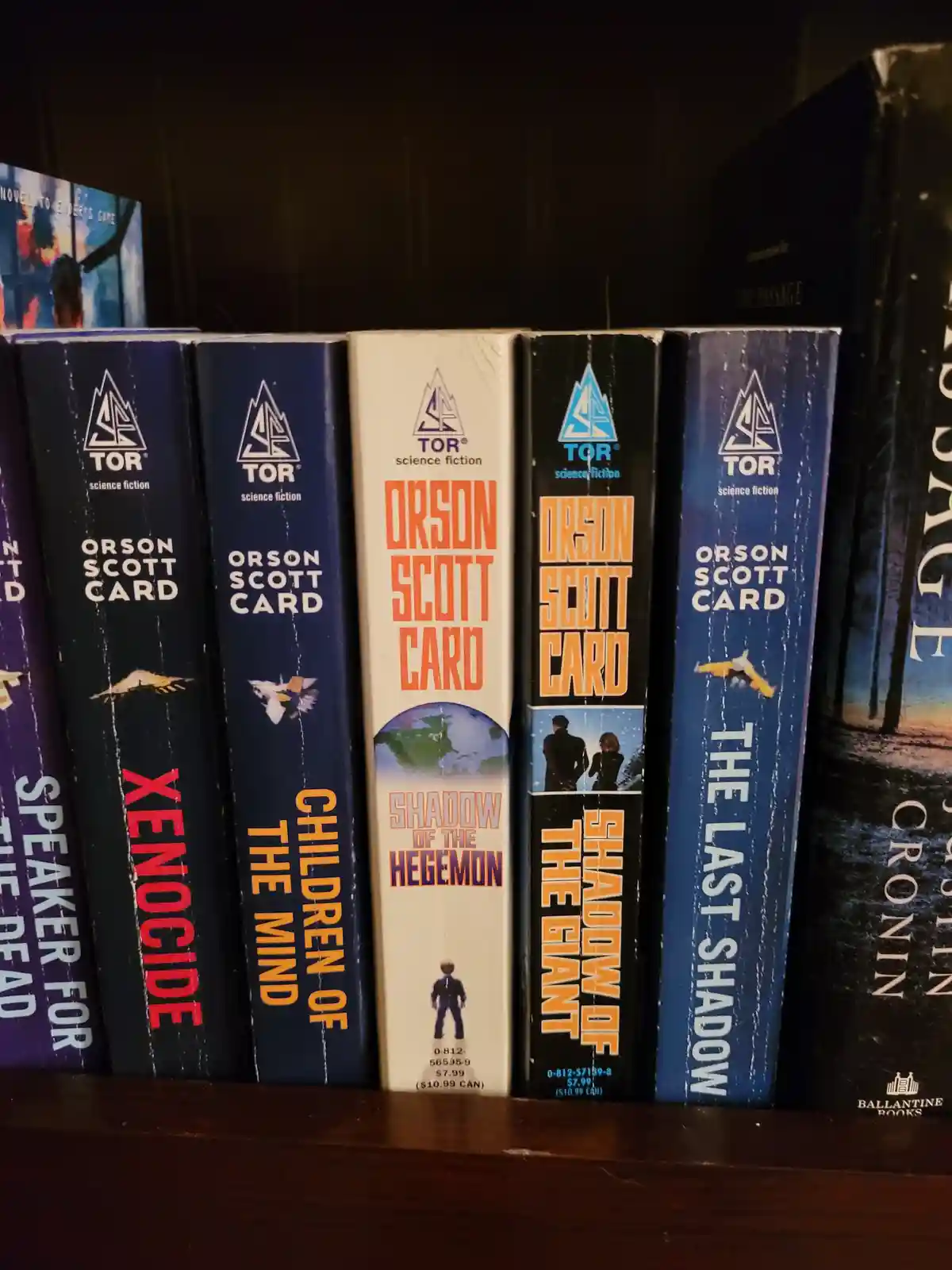 A close-up of several science fiction books by Orson Scott Card on a dark wooden shelf, showing spines with titles like…