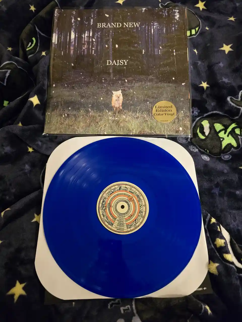 A blue vinyl record from the band Brand New, titled 'Daisy', is displayed with its album cover. The cover features a photo…
