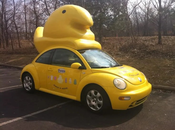 A bright yellow Volkswagen Beetle is parked in a lot with a giant, oversized yellow Peep marshmallow candy figure perched on…