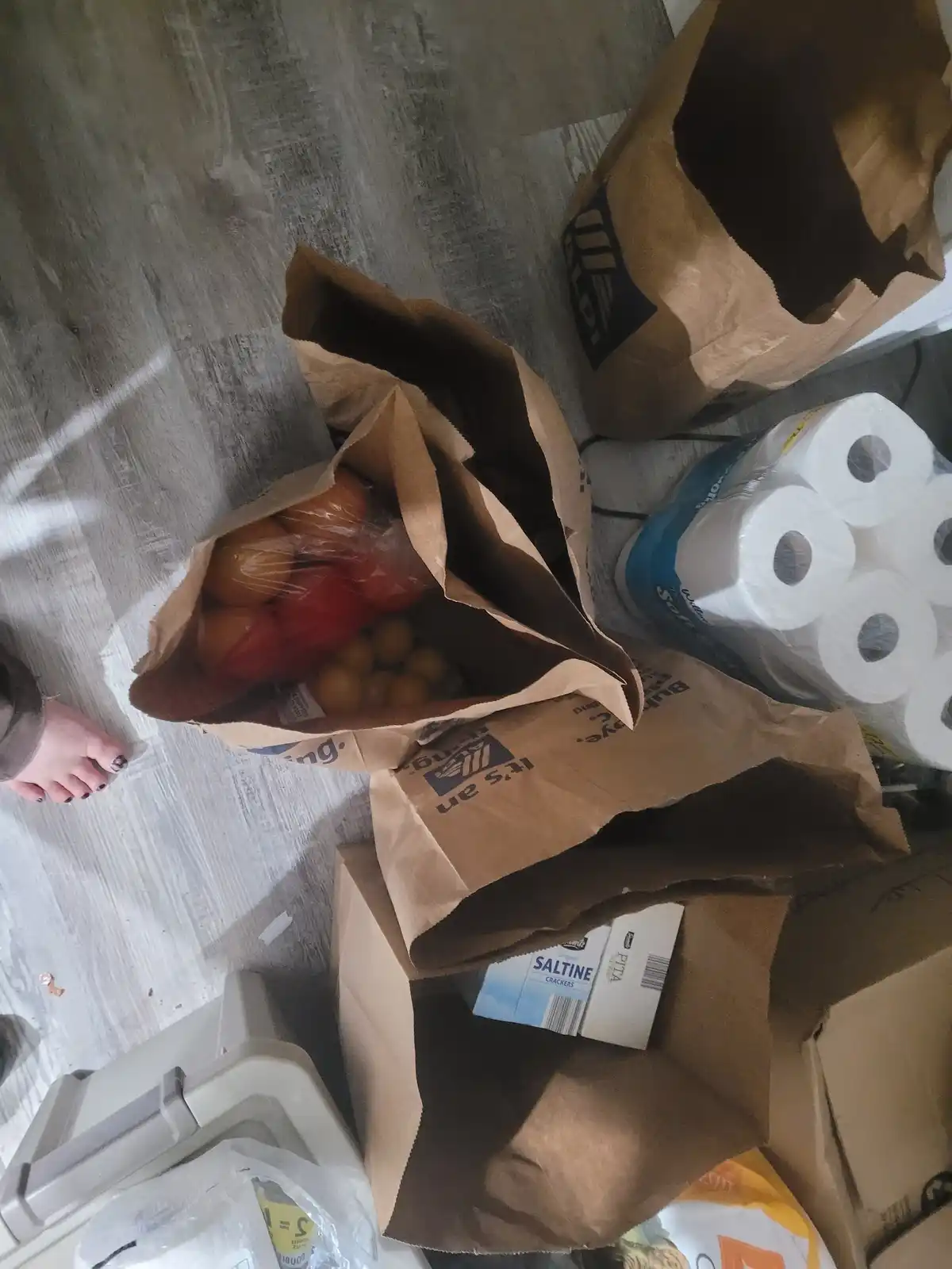 A top-down view of grocery bags on a wooden floor, one open bag reveals tomatoes and small fruits, another contains saltine…