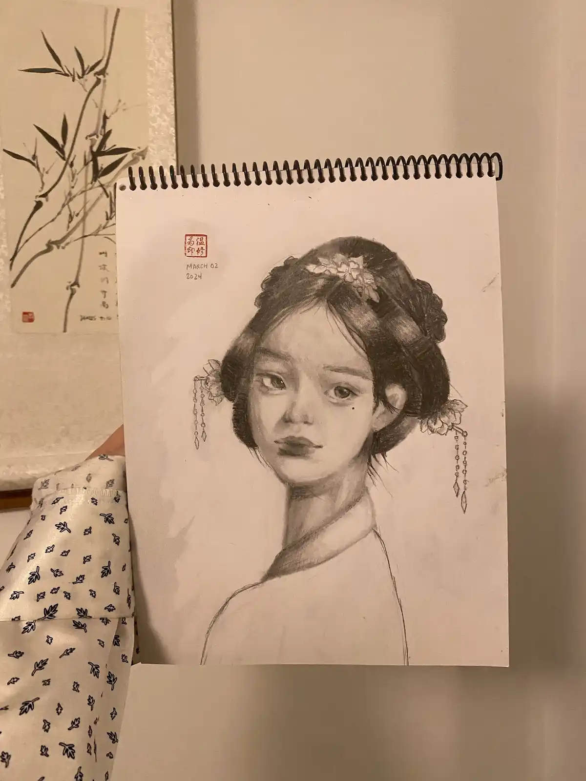 A person holds up a spiral-bound sketchbook displaying a detailed pencil drawing of a woman with traditional Chinese…