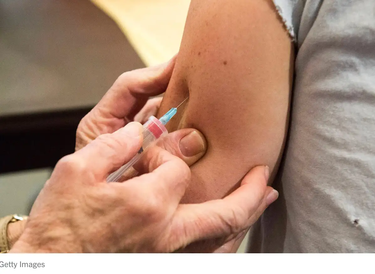 A close-up photo shows a person's arm being injected with a syringe by another person's hands. The recipient wears a grey…