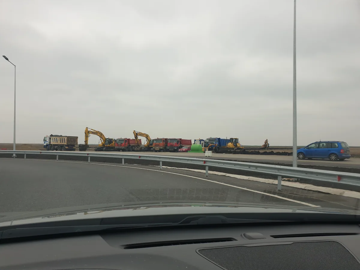 A photo taken from inside a car shows a line of heavy construction vehicles, including excavators and dump trucks, parked…