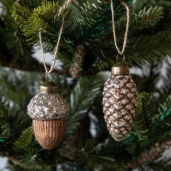 Two glittery Christmas ornaments shaped like an acorn and a pinecone hang from twine on a green pine tree branch. The acorn…