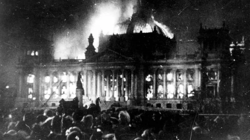 A historical black-and-white photograph shows a large, ornate building engulfed in flames at night, with crowds of people…