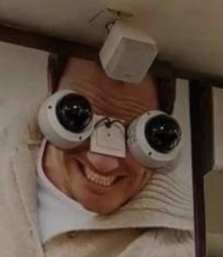 A man with a wide, toothy grin has two white security cameras mounted over his eyes, with a small white tag hanging between…