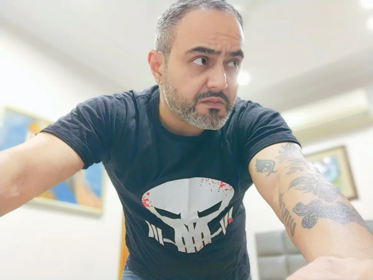 A man with a grey beard and tattoos on his arms wears a black t-shirt with a white skull logo and looks sideways with a…