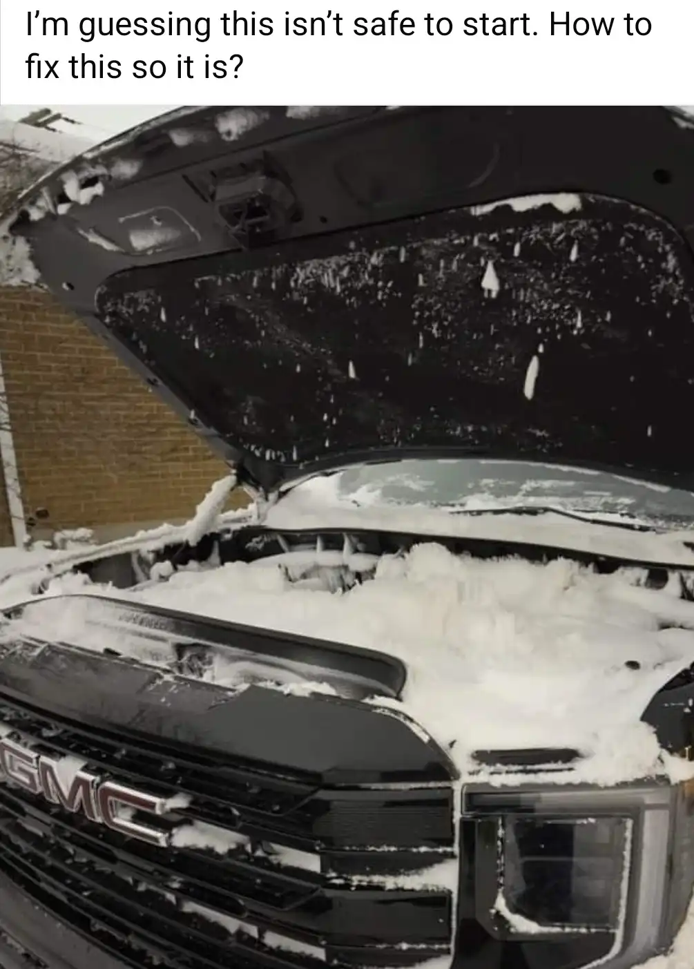 A black GMC truck's hood is open, revealing an engine compartment completely buried under thick, fluffy snow. Snow also…