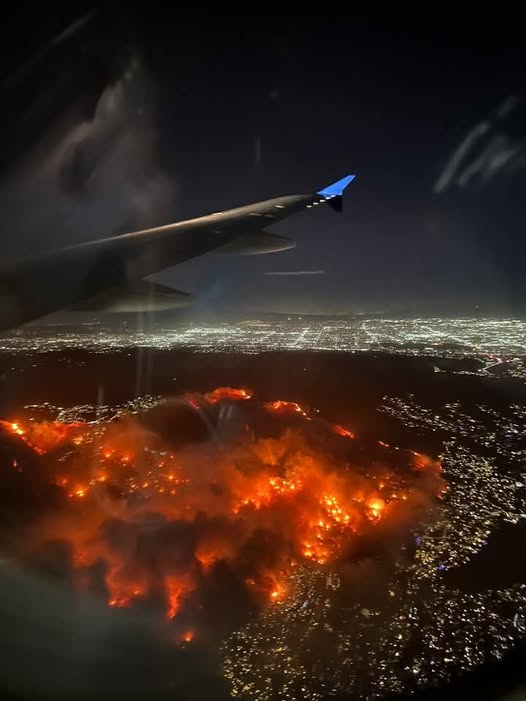An aerial photo taken from an airplane window shows a large, glowing orange fire spreading across a landscape at night, with…