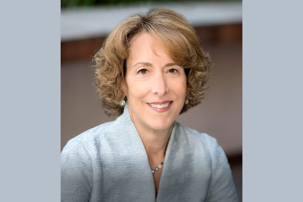 a professional headshot of a smiling woman with short, wavy brown hair, wearing a light blue textured blazer and pearl…