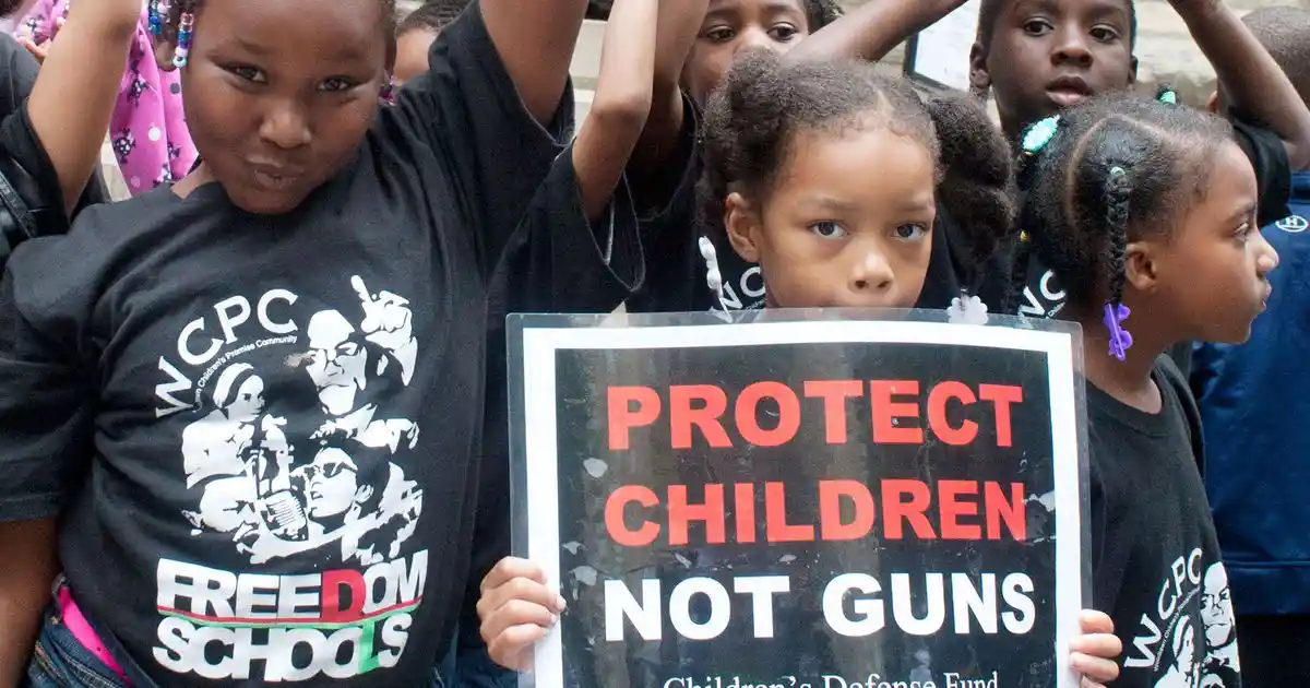 A group of young Black children, some wearing black t-shirts with 'WCPC FREEDOM SCHOOLS' graphics, are gathered outdoors…