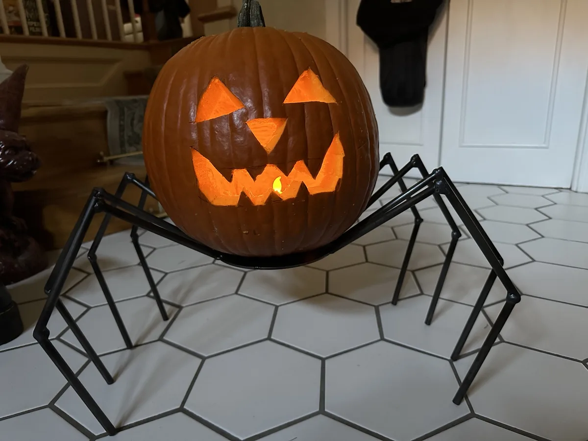 A carved jack-o'-lantern with a glowing face sits atop a black metal spider stand on a white hexagonal tiled floor. The…