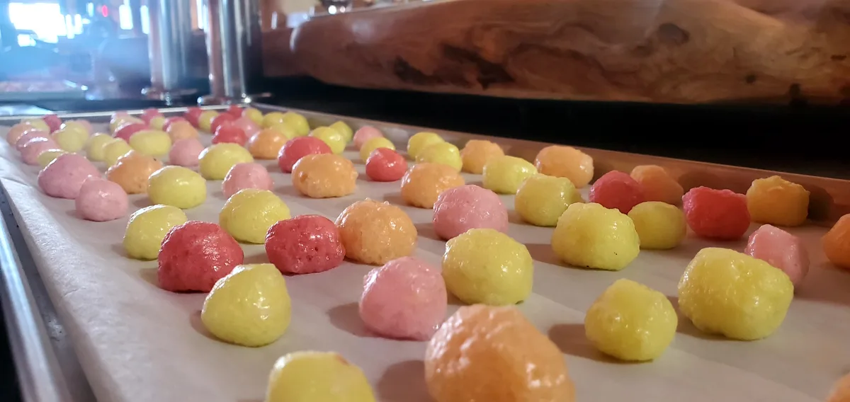A baking sheet lined with parchment paper holds dozens of small, round, colorful balls in pink, yellow, and orange…