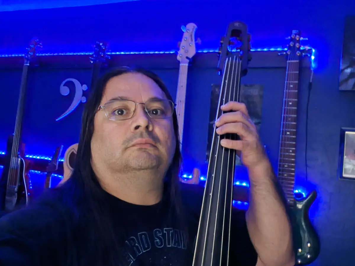 A man with long dark hair and glasses holds a double bass in a room lit with blue lights, surrounded by other bass guitars…
