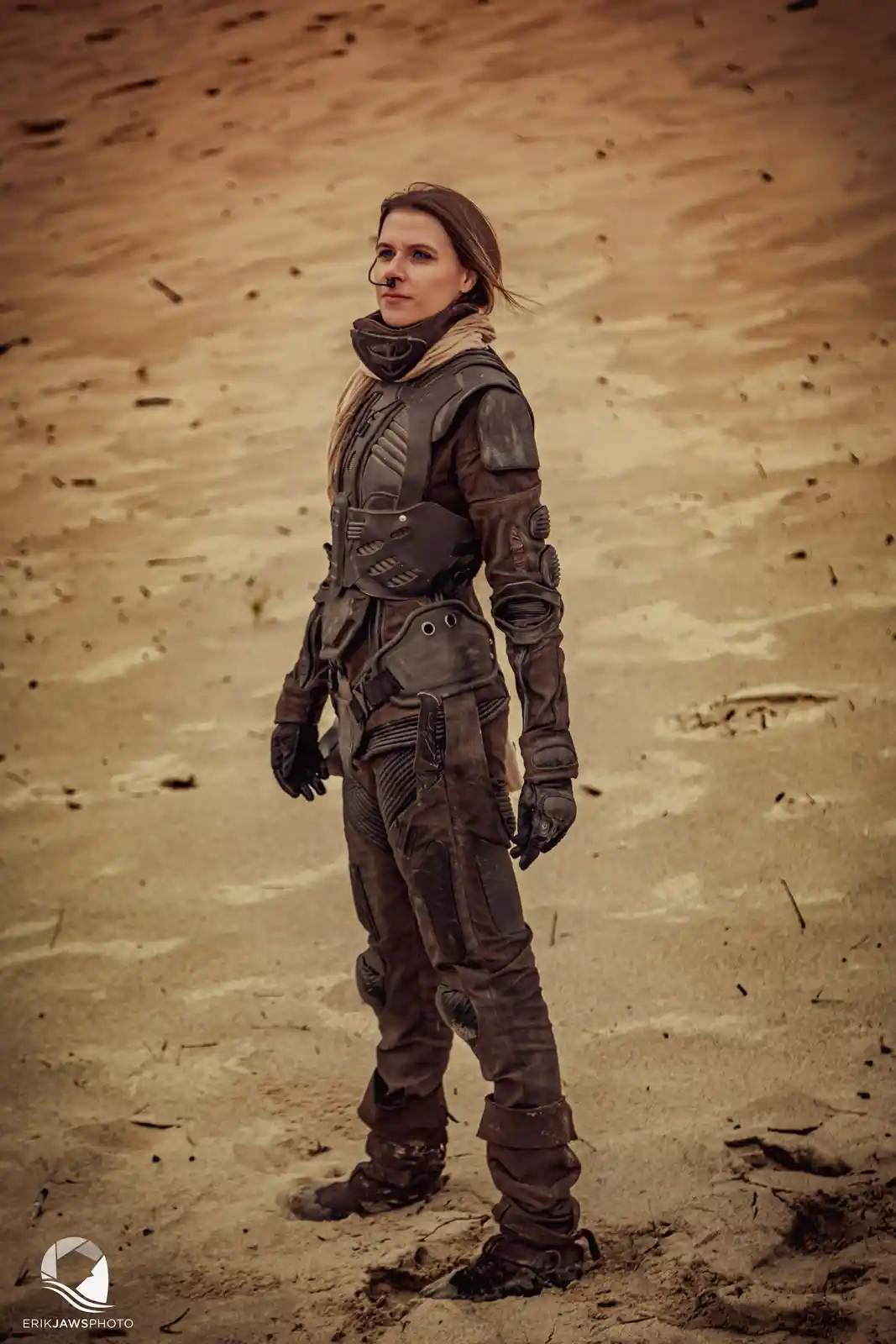 A woman in a full-body, dark, futuristic desert survival suit stands in a sandy, barren landscape. She has a small breathing…