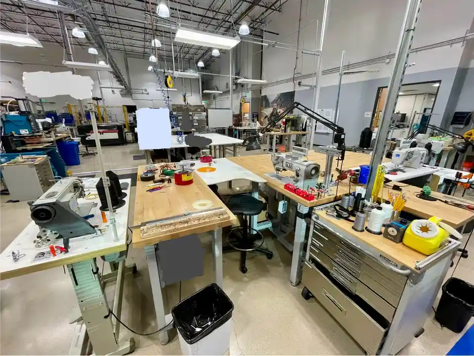 A wide shot of a well-lit, spacious workshop filled with multiple workstations, sewing machines, tools, spools of thread…