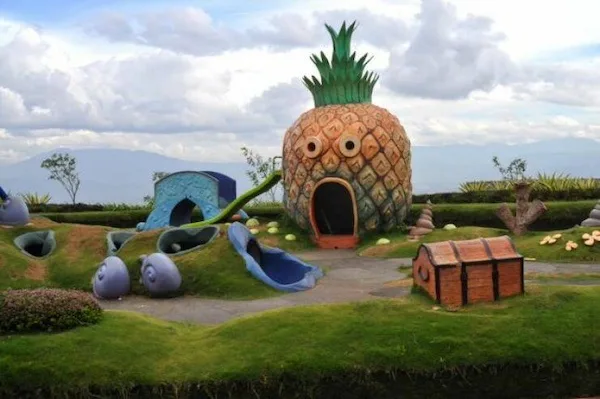 A large, realistic pineapple-shaped house with cartoonish eyes and a mouth, set in a landscaped garden with other whimsical…