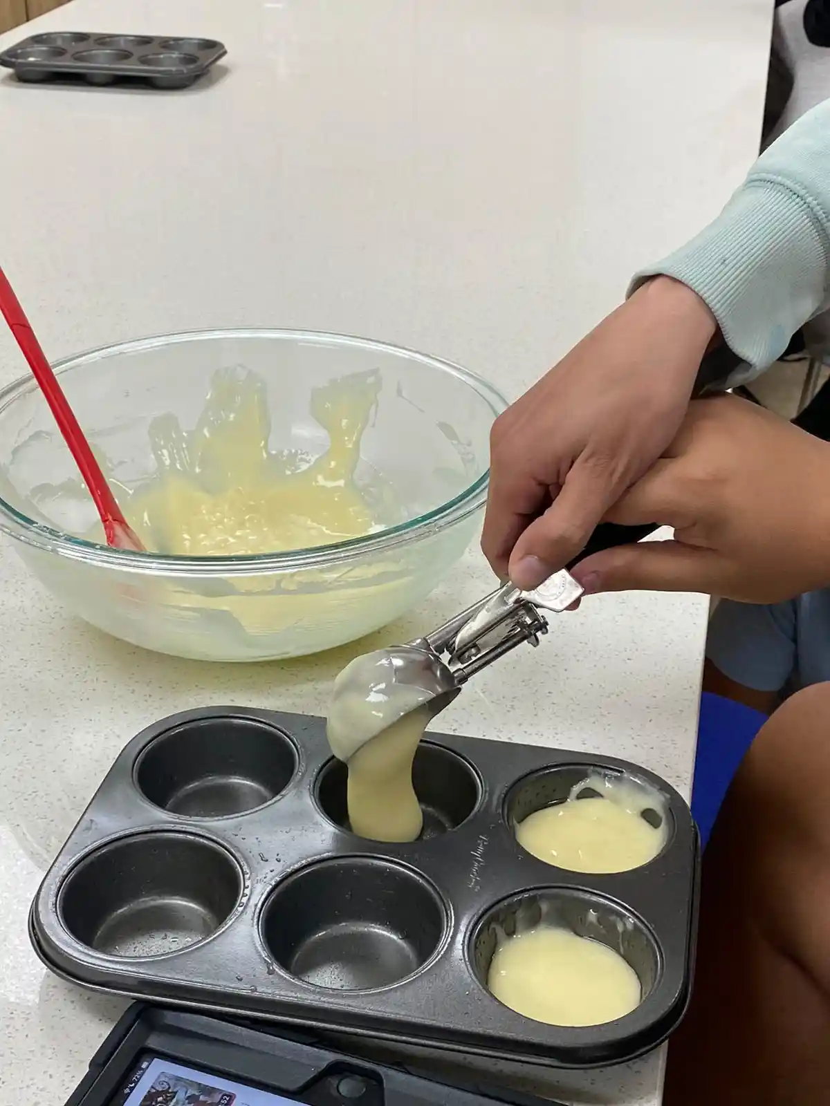 A person is using a metal cookie scoop to portion yellow batter into a dark muffin tin, with a glass mixing bowl containing…