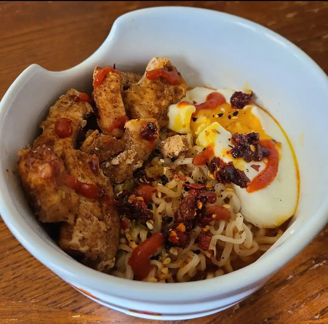 a close-up photo of a white bowl filled with ramen noodles, fried chicken pieces, and a soft-boiled egg, all topped with red…