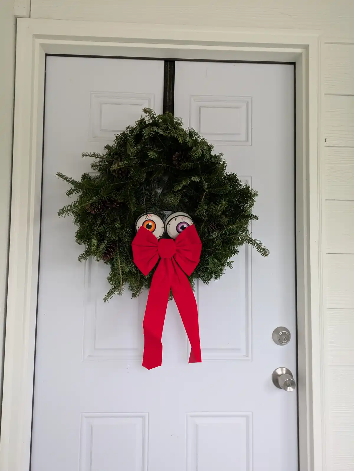 A festive green wreath with pinecones hangs on a white door, featuring two large, cartoonish eyeballs for eyes and a bright…