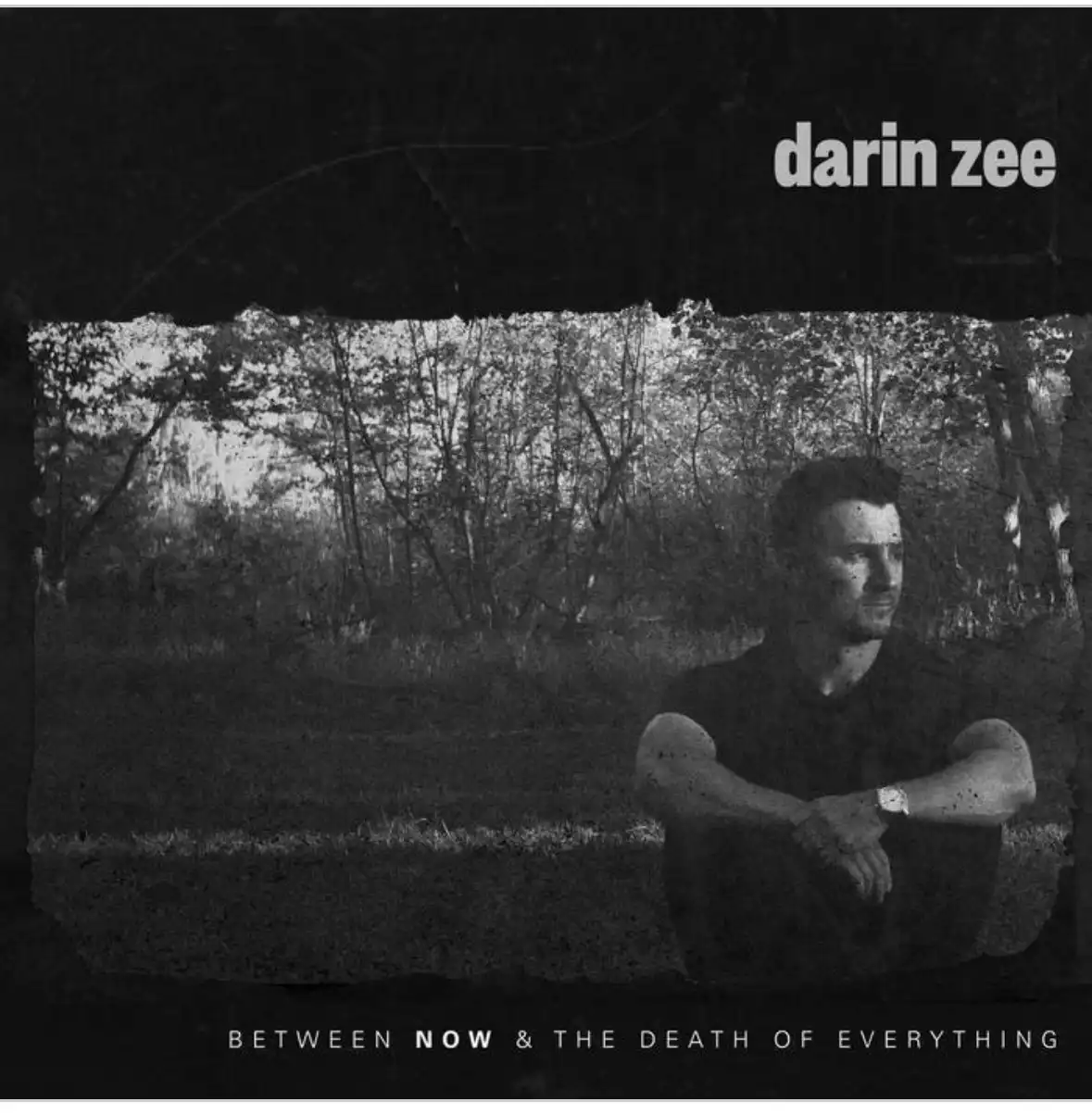A black-and-white album cover featuring a man sitting outdoors in a wooded area, looking pensive. The text 'darin zee'…