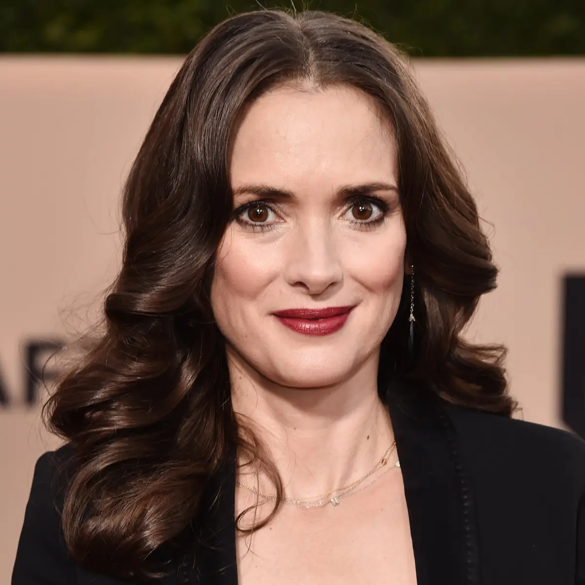 A close-up photo of actress Winona Ryder smiling at the camera, wearing a black blazer, layered necklaces, and dangling…