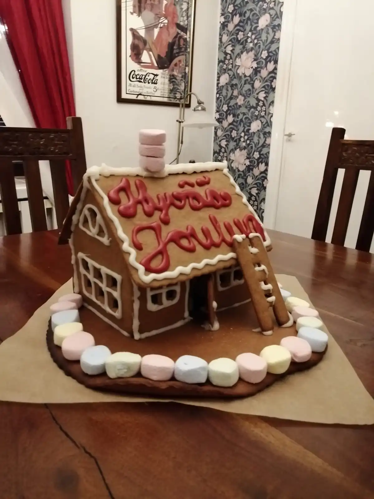 A gingerbread house decorated with white icing and colorful marshmallows sits on a wooden table. The roof features red icing…