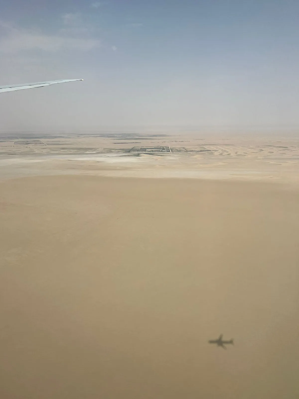 An aerial view from an airplane window shows the wing in the top left corner and the shadow of the plane cast on a vast…