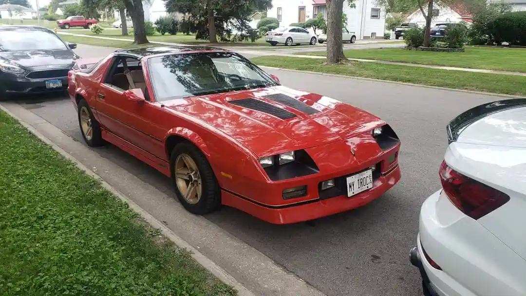A red 1980s Pontiac Firebird Trans Am with black hood stripes and gold wheels is parked on a residential street, flanked by…