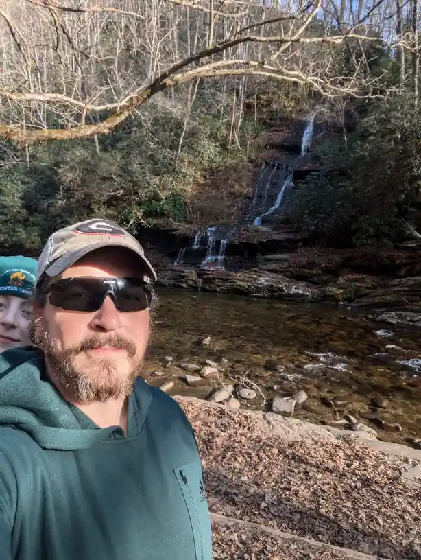 A selfie of a man with a beard and sunglasses wearing a green hoodie and a baseball cap, standing in front of a small…
