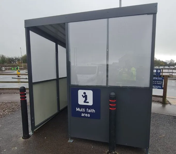An outdoor shelter with translucent panels and a blue sign reading 'Multi faith area' with a pictogram of a person. The…