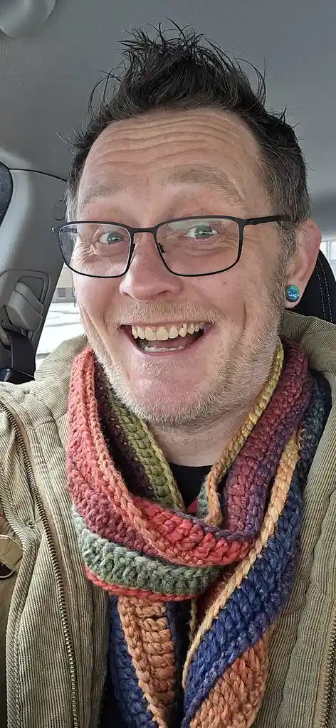 A man with glasses and a colorful, multi-strand crocheted scarf smiles broadly while sitting in a car. The scarf features…