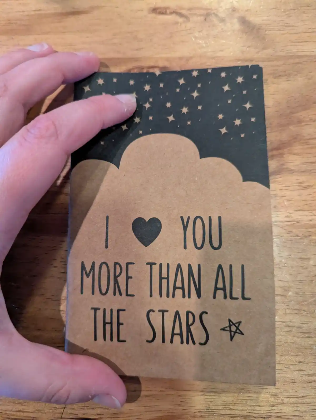 A hand holds a brown paper card with a black sky and star pattern above a cloud shape. The card reads 'I ❤️ YOU MORE THAN…