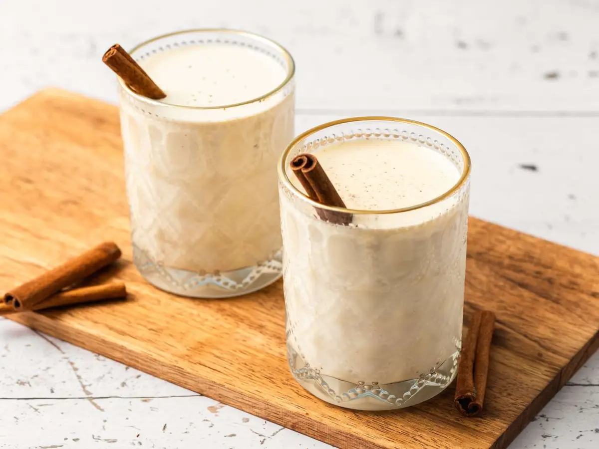Two glasses of creamy eggnog, each garnished with a cinnamon stick, sit on a wooden cutting board. Additional cinnamon…