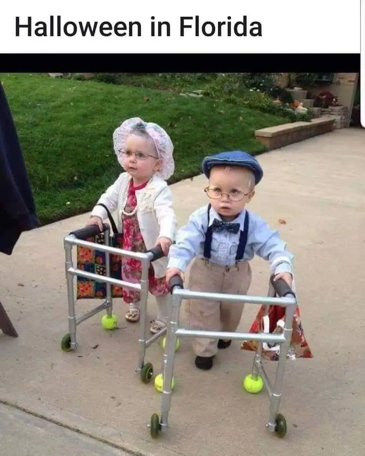 Two young children, a boy and a girl, are dressed in vintage-style outfits and wearing glasses, each using a wheeled walker…
