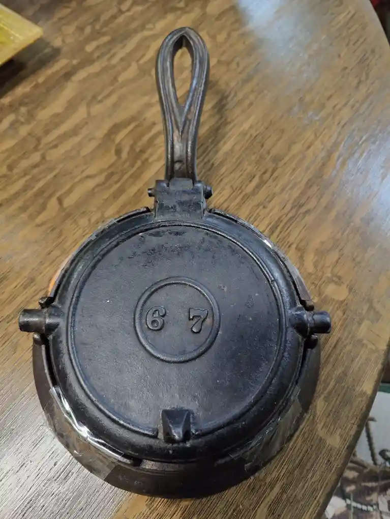 A black cast iron waffle iron sits closed on a wooden table, showing the numbers '6' and '7' embossed in a circle on its…
