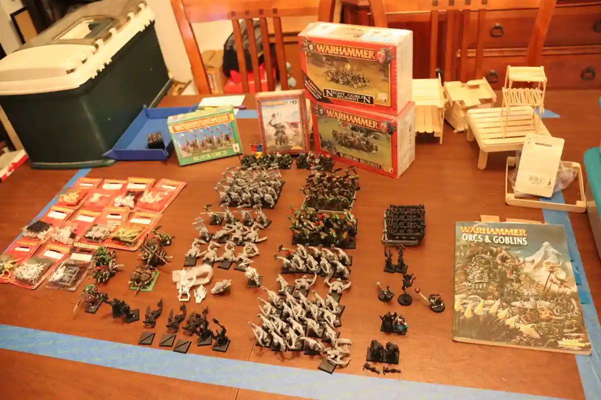 A wooden table is covered with Warhammer fantasy miniatures, unpainted and painted figures, boxed sets labeled 'Night…