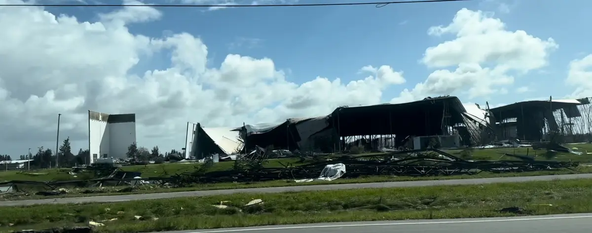 A wide shot shows a large industrial building with its roof and walls completely torn off, exposing twisted metal beams and…