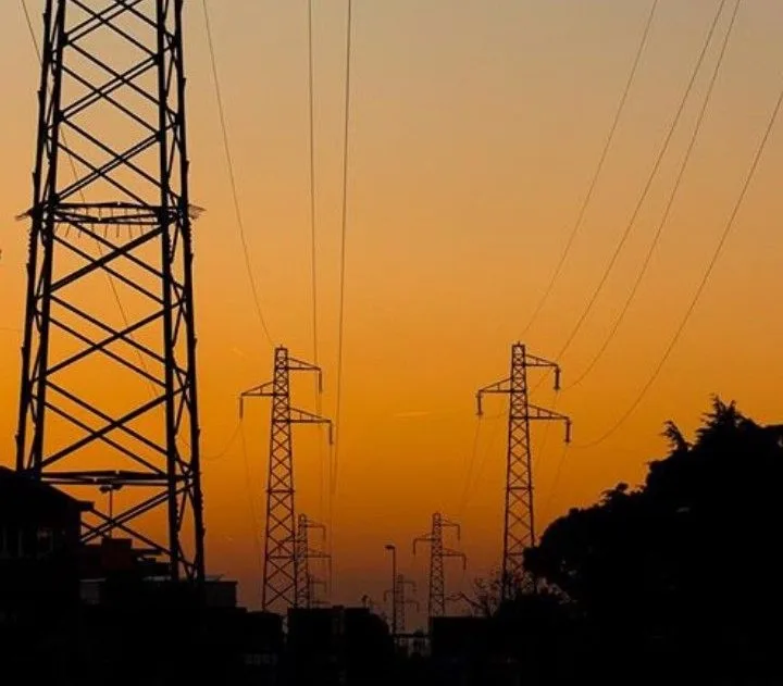 A silhouette of multiple electricity pylons against a vibrant orange sunset sky, with dark trees and buildings at the…