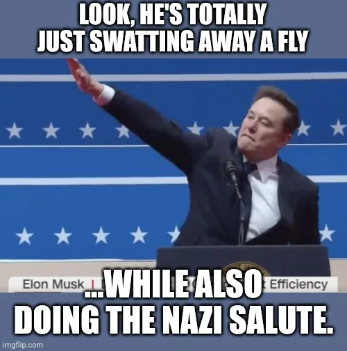A man in a dark suit stands at a podium with a blue background featuring white stars and stripes. He raises his right arm in…