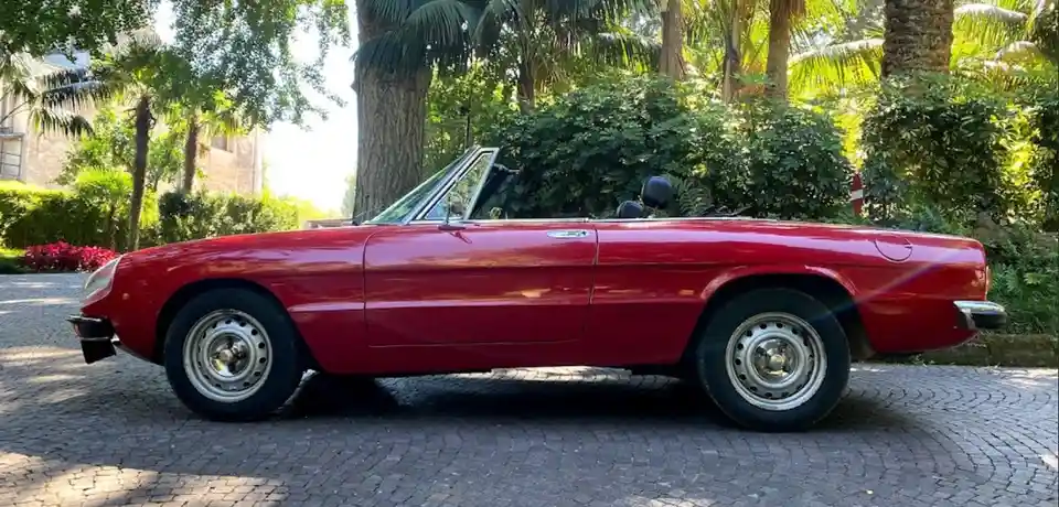 a red alfa romeo spider convertible is parked on a cobblestone driveway surrounded by lush greenery and palm trees. The…