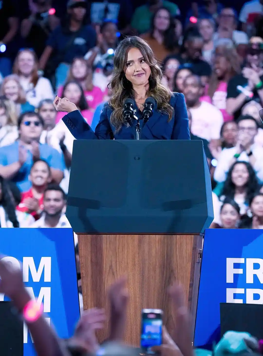 A woman in a dark blue blazer stands at a wooden podium with microphones, smiling smugly while raising her fist. Behind her…
