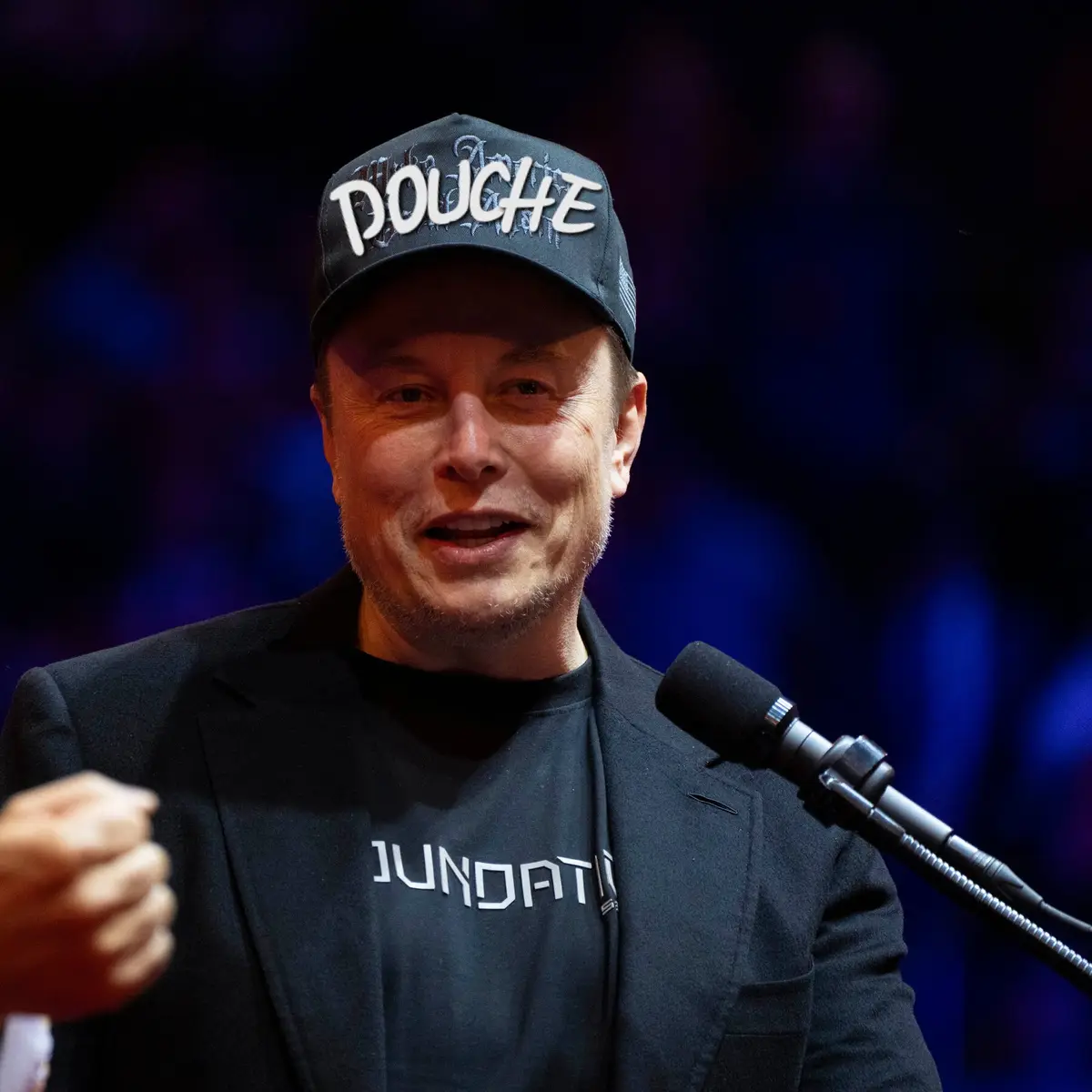 A man wearing a black cap with the word 'DOUCHE' in white graffiti-style lettering and a black t-shirt with 'JUNDATI'…