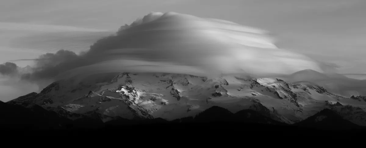 A black-and-white photograph shows a snow-covered mountain range under a dramatic, flowing cloud formation that resembles a…
