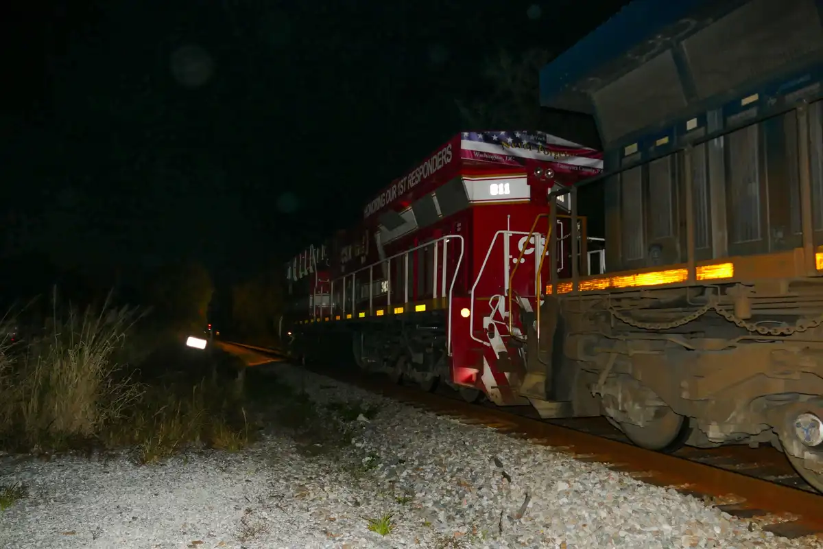 A red train engine at night with "HONORING OUR 1ST RESPONDERS" written on its side, alongside "Never Forget" and location…