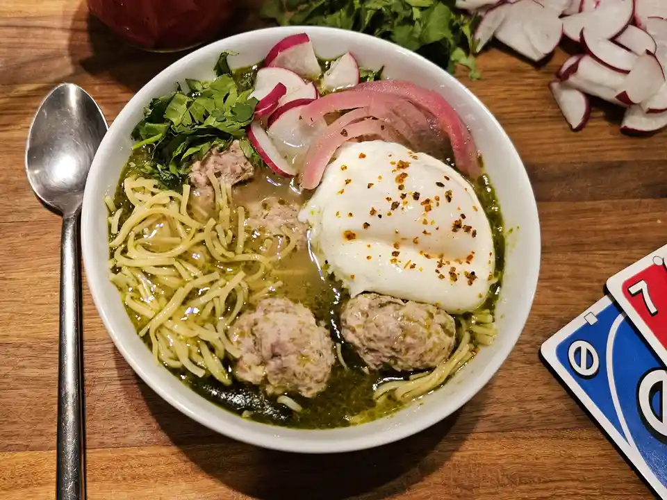 A bowl of pasta with meatballs, a poached egg, radishes, and herbs sits on a wooden table next to a spoon and Uno cards. The…