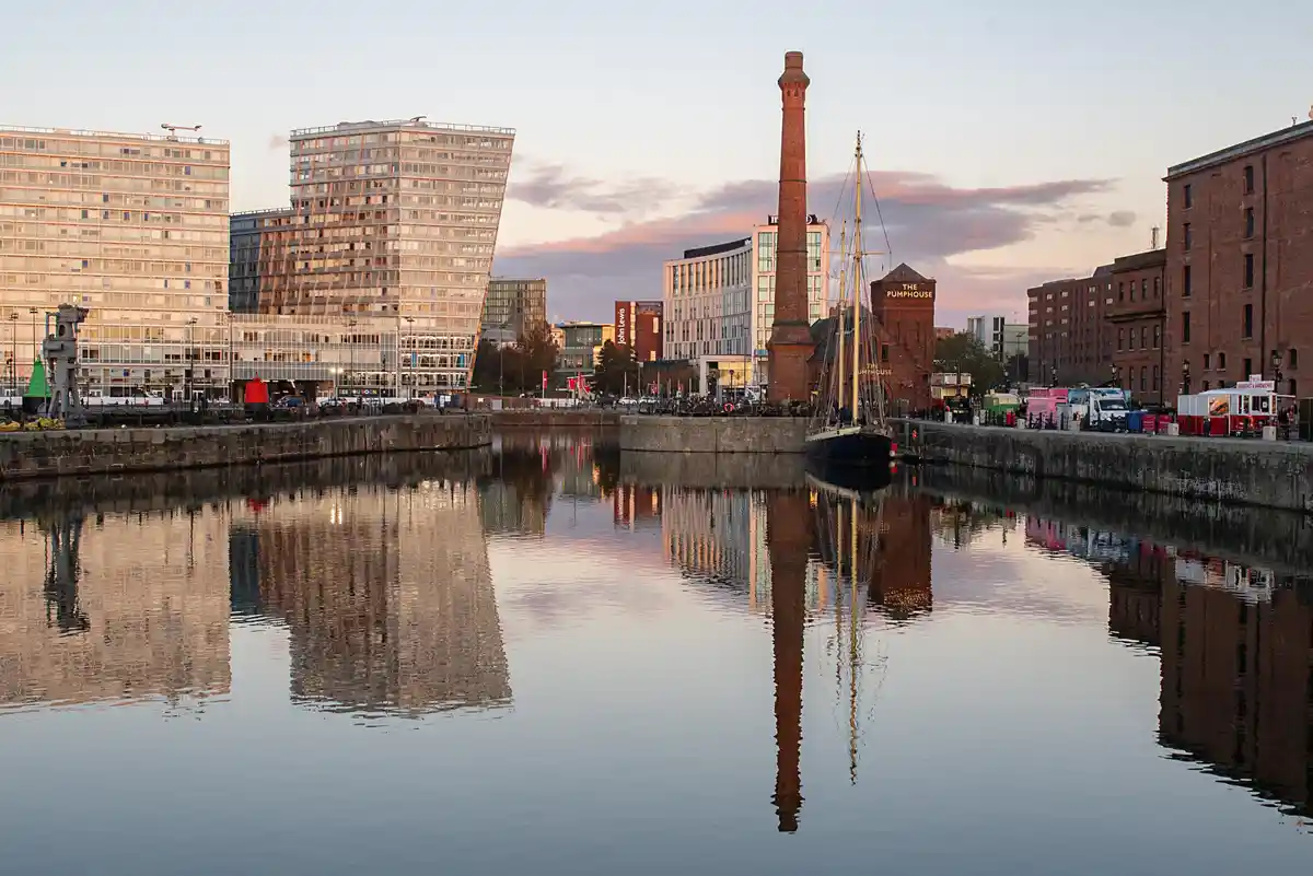 A serene waterfront scene at dusk with modern glass buildings and historic brick structures reflected in calm water…