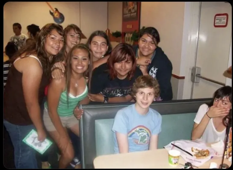A group of young people are posing for a photo in a diner booth. Most are smiling and leaning in, but one boy in a blue…