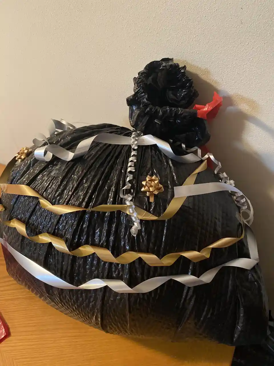 A large, overstuffed black trash bag is decorated with shiny gold and silver ribbons and bows, sitting on a wooden surface…